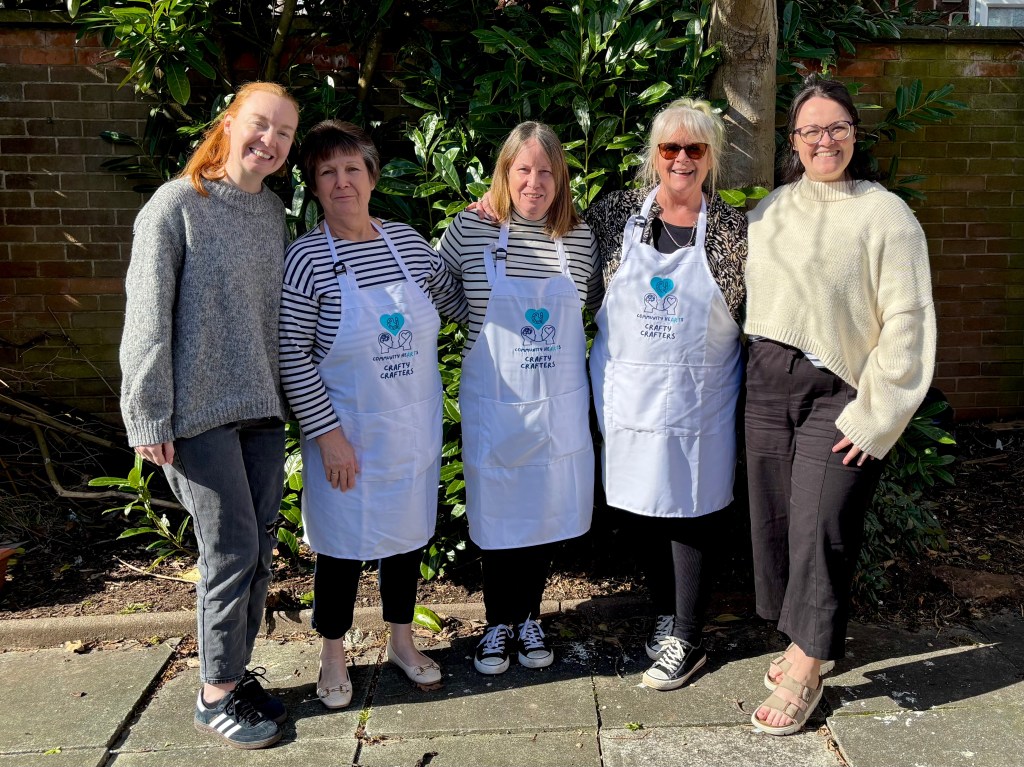  The Board of Trustees - A group of five women standing together outdoors, wearing white aprons with the Community HeARTs logo, smiling for the camera in front of green foliage.